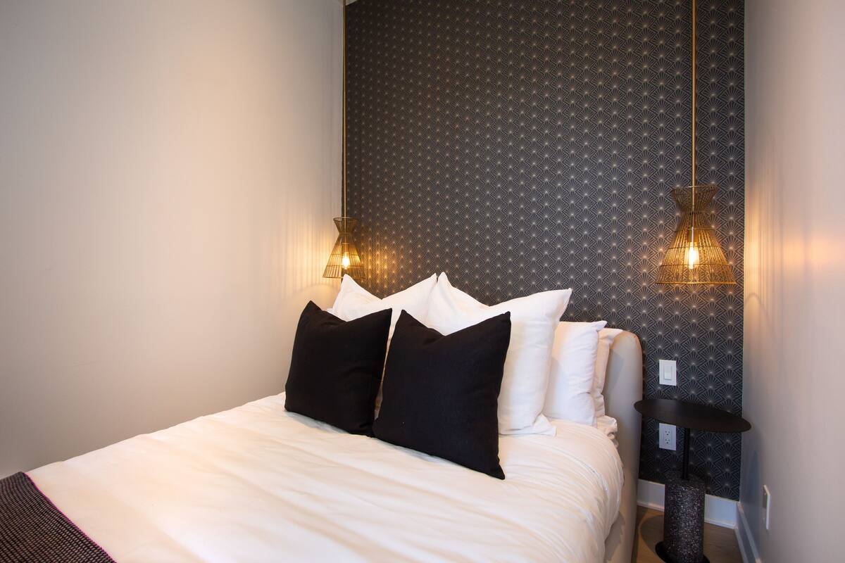A neatly made bed with white linens and black pillows, a patterned wallpaper, and two wall-mounted lamps.