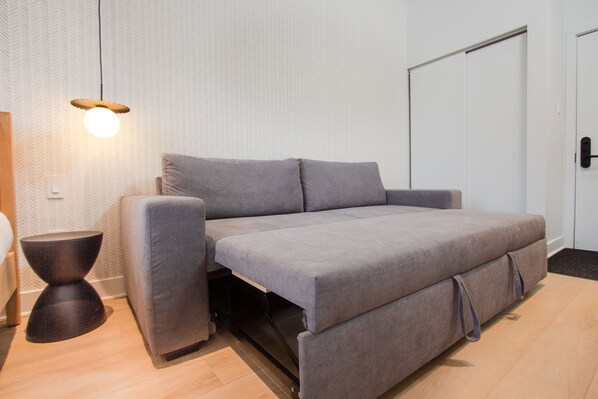 Une chambre moderne avec un canapé-lit gris, une petite table d’appoint ronde et un luminaire suspendu.