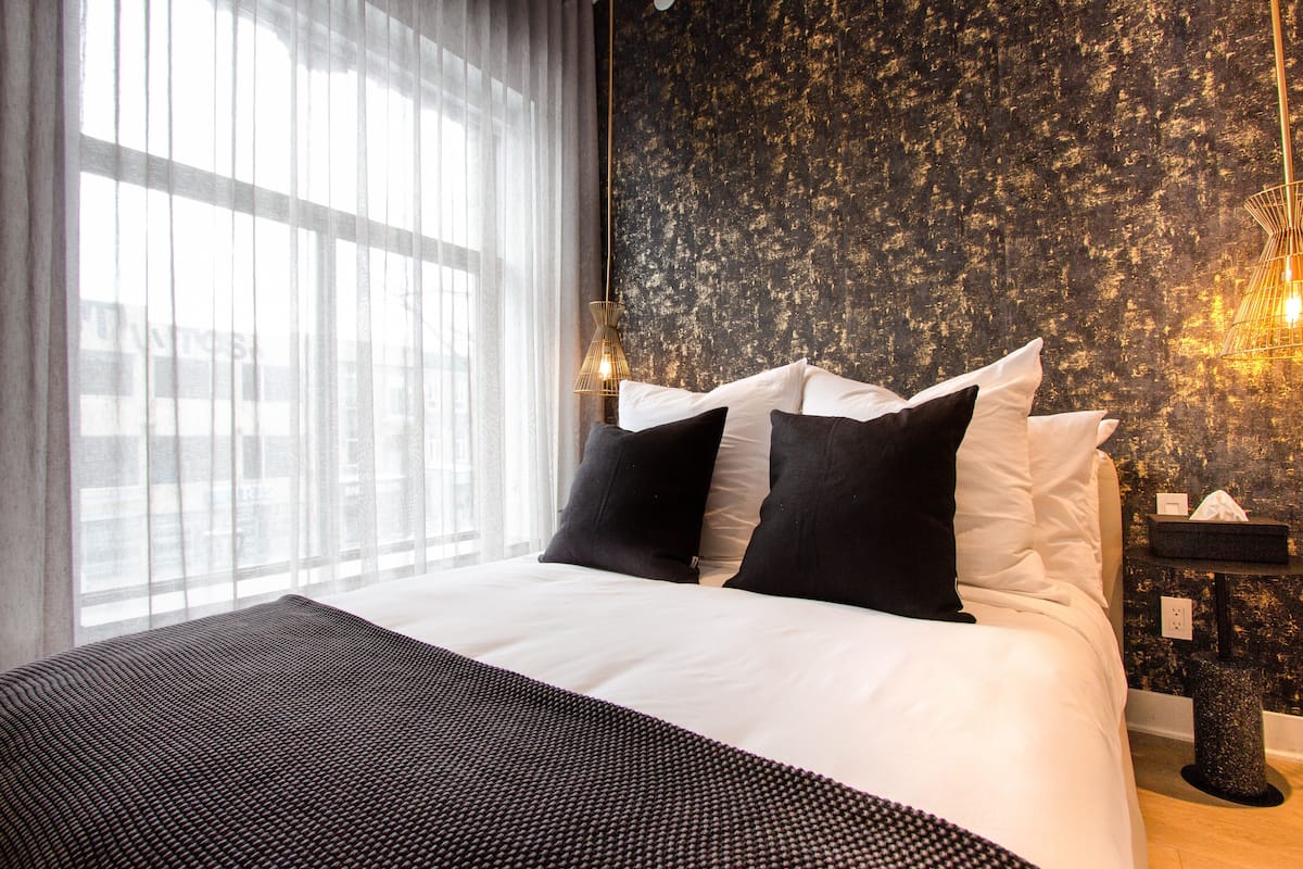 A neatly made bed with white and black bedding, a textured wall behind, and a window with sheer curtains.