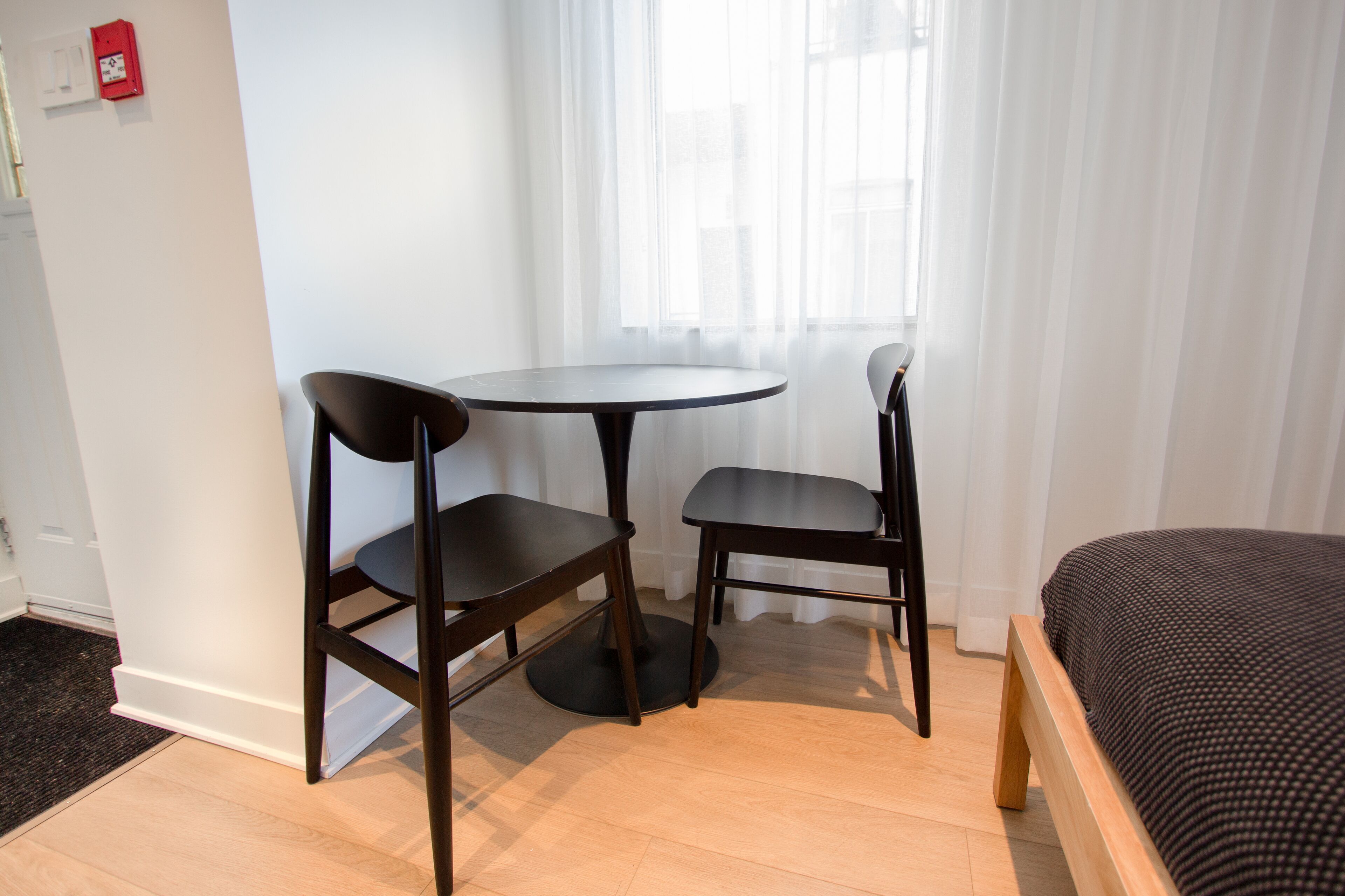 Una habitación con cama, una mesa redonda, dos sillas y un ventanal con cortinas translúcidas.