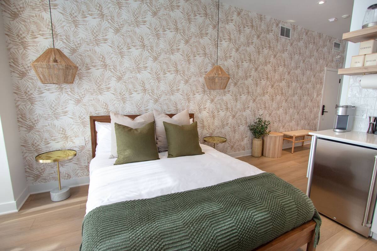 A modern hotel room with a large bed, two bedside tables, a small desk, a kitchenette, and a patterned wallpaper.