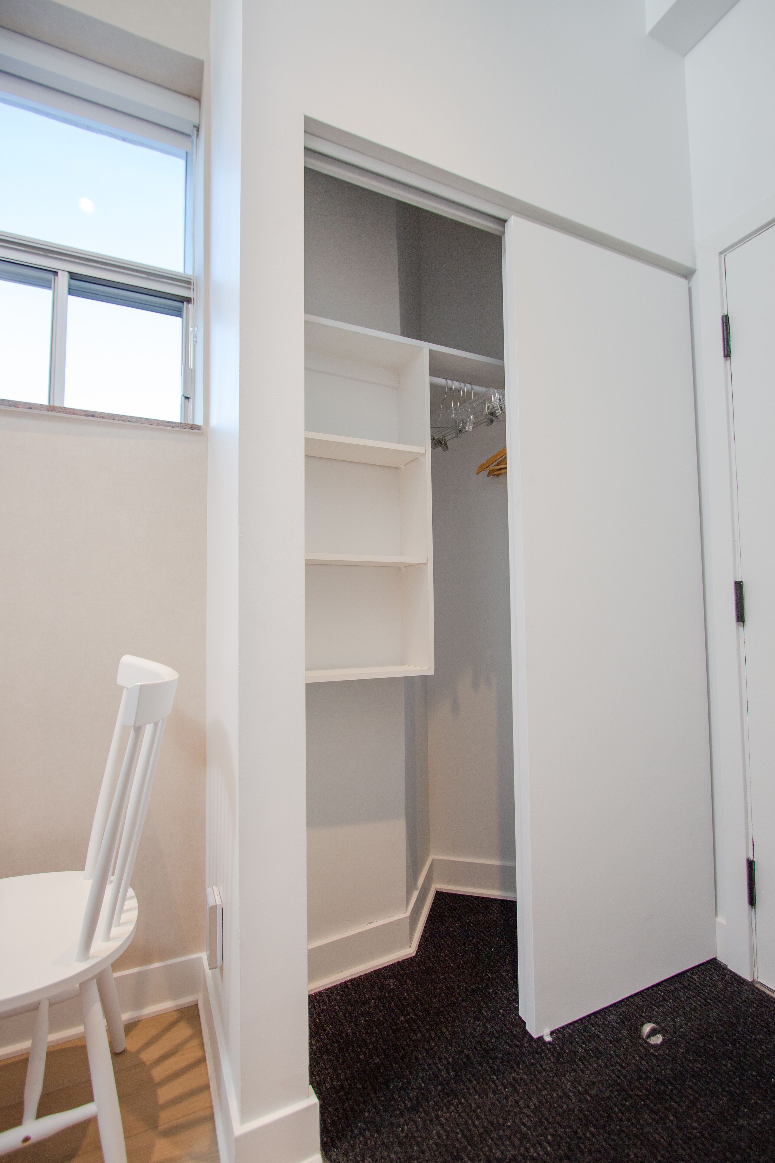 A room with a white chair, a closet with shelves, and a window.