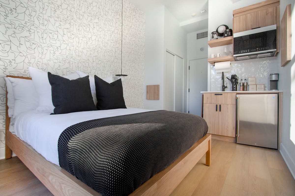 A modern bedroom with a large bed, a wooden headboard, and a kitchen area with a refrigerator and microwave.