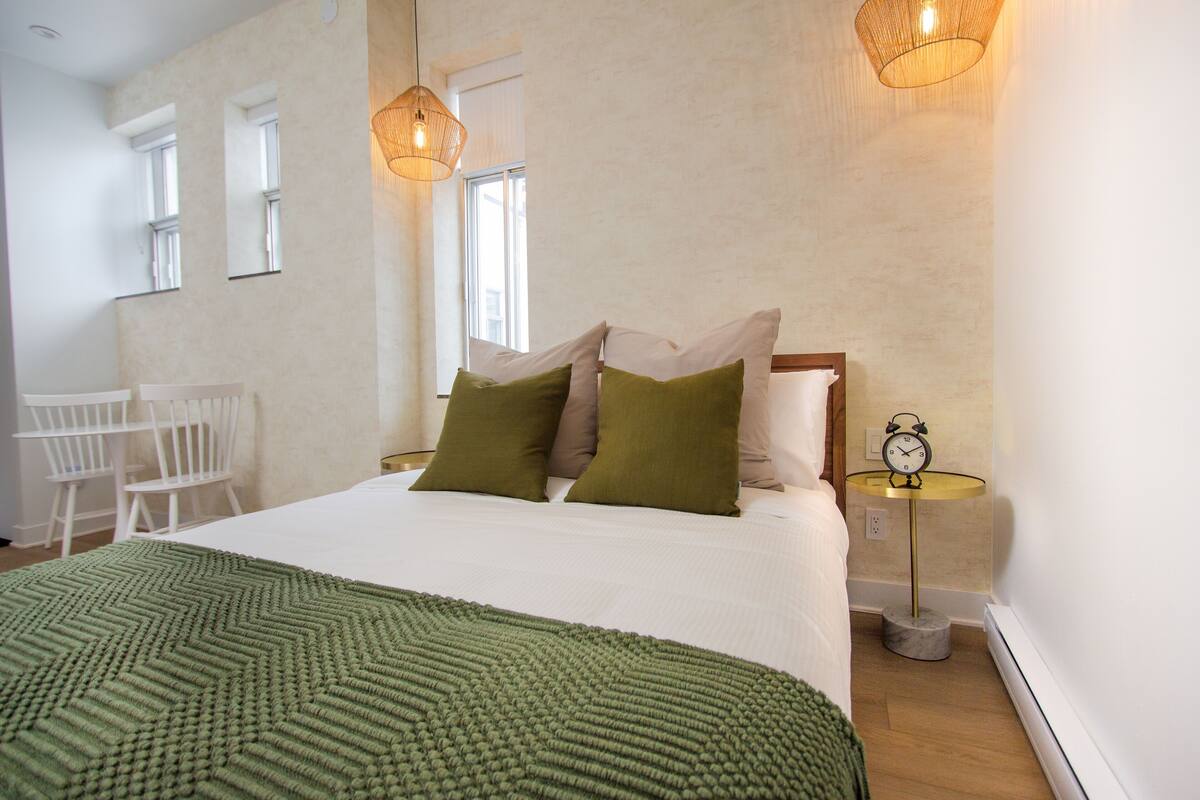 A neatly made bed with a green blanket, two pillows, and a bedside table with a clock.