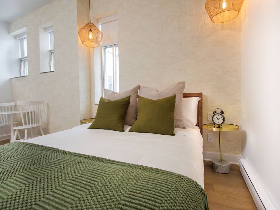 Un lit bien fait, recouvert d’une couverture verte, avec deux oreillers et une table de chevet sur laquelle se trouve une horloge.
