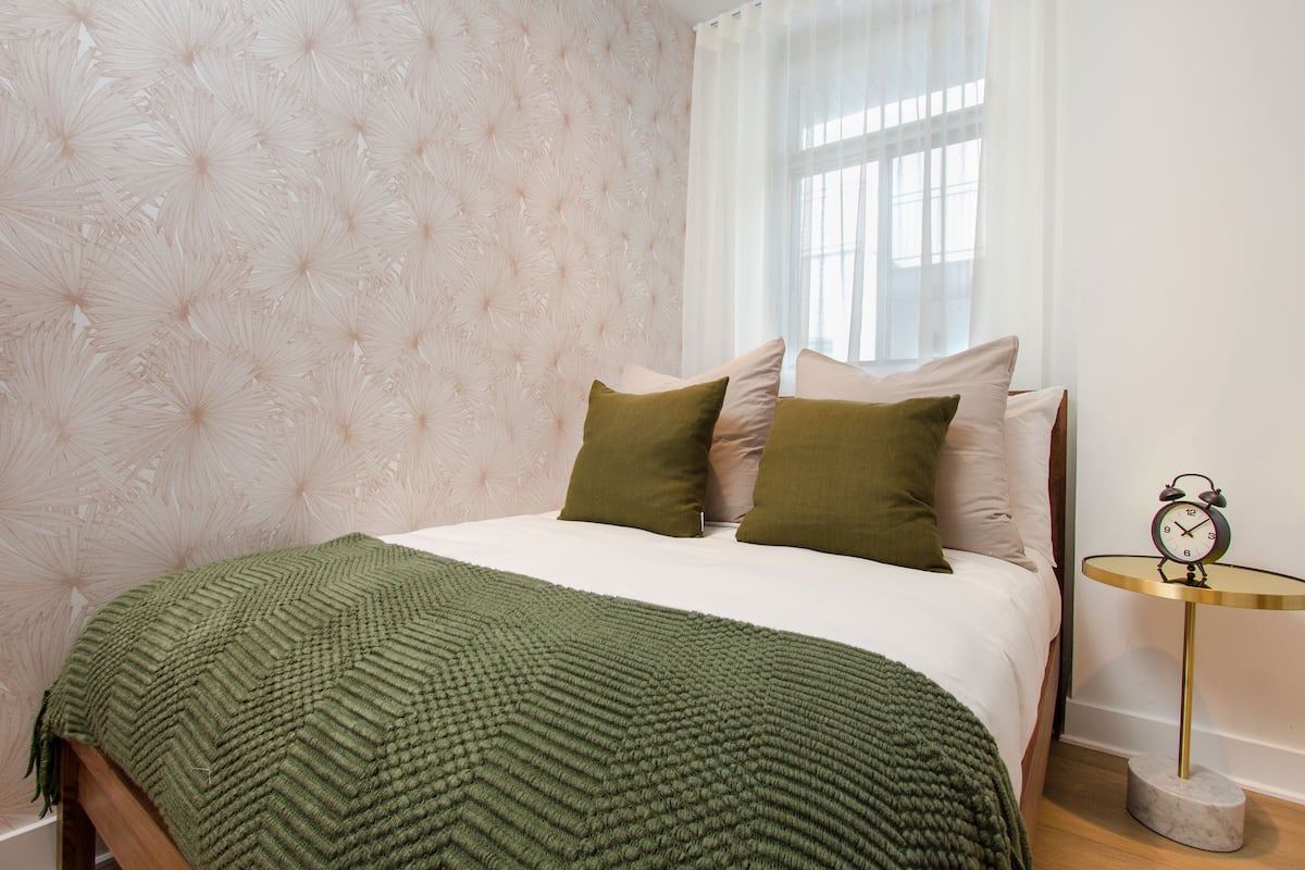 A neatly made bed with a green blanket, a tufted wall, and a small bedside table with a clock.