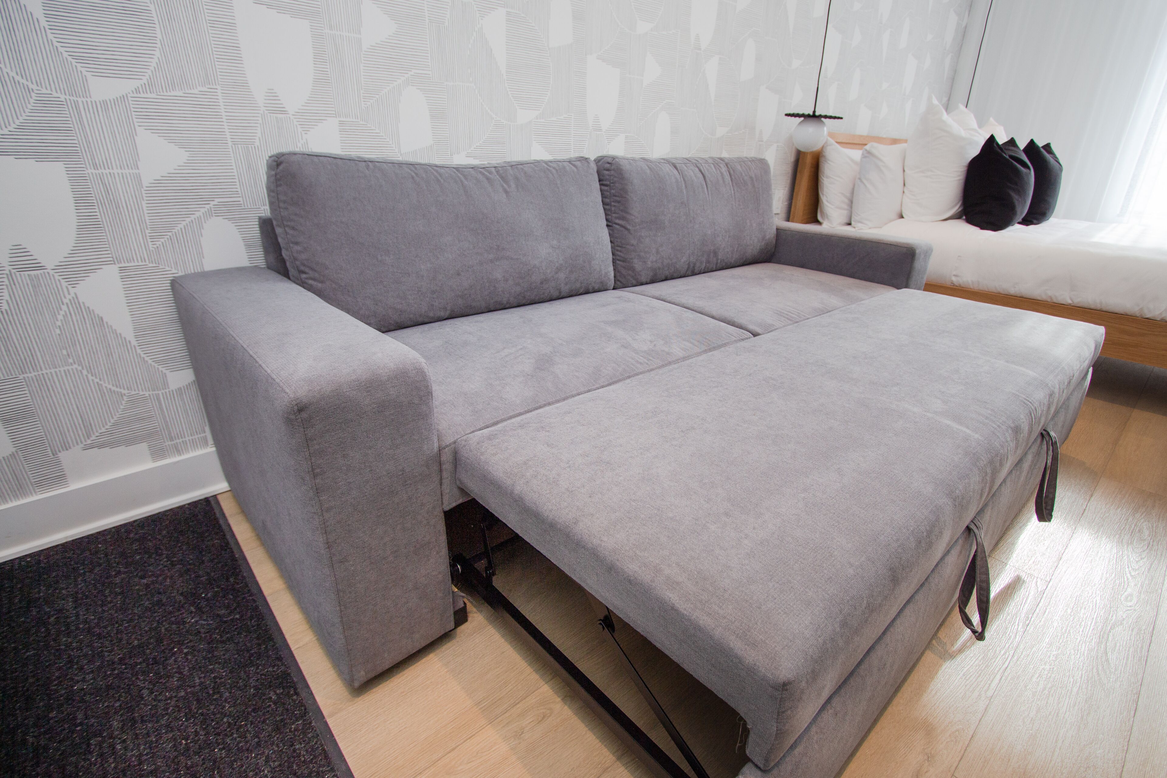 A grey sofa with a wooden frame, a white wall with a geometric pattern, and a small side table with a lamp.