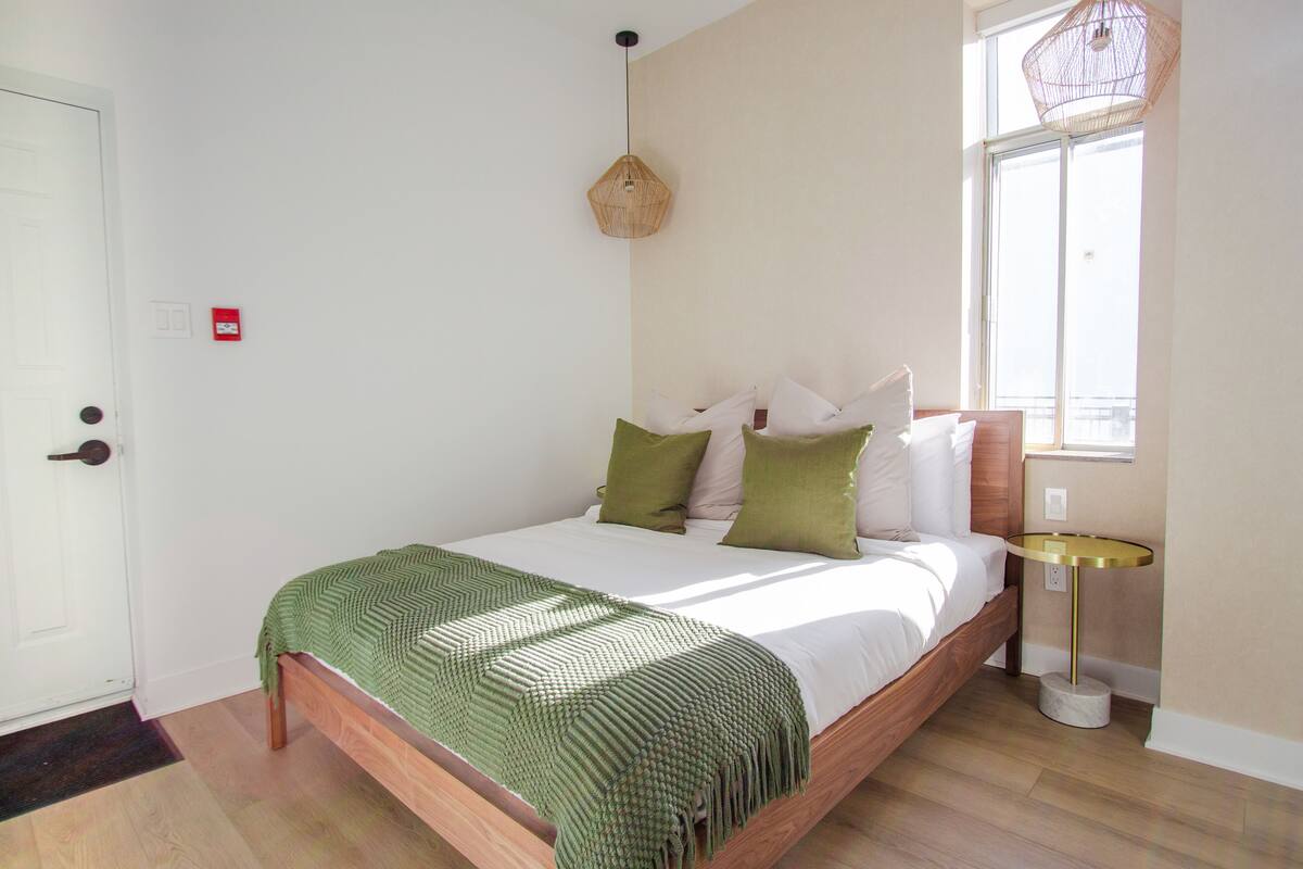 A bedroom with a wooden bed, green throw, and a round table with a lamp.