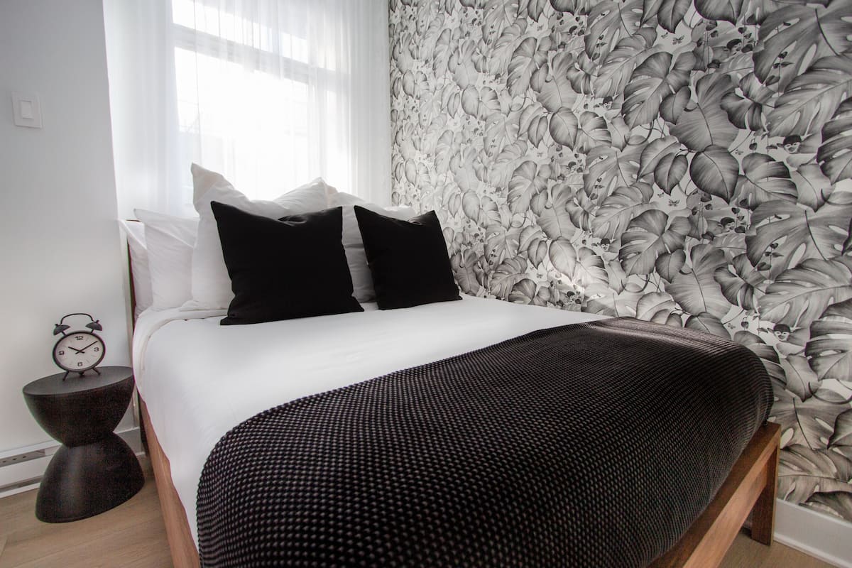 A neatly made bed with a white duvet and black pillows, a dark wooden bedside table with a clock, and a wall with a floral wallpaper pattern.