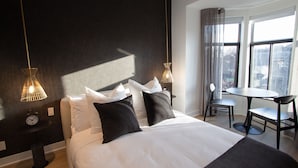 A hotel room with a large bed, two bedside tables, a desk with a chair, and a view of the city through large windows. - Unique Stays - 4119 St-Denis (Montreal)