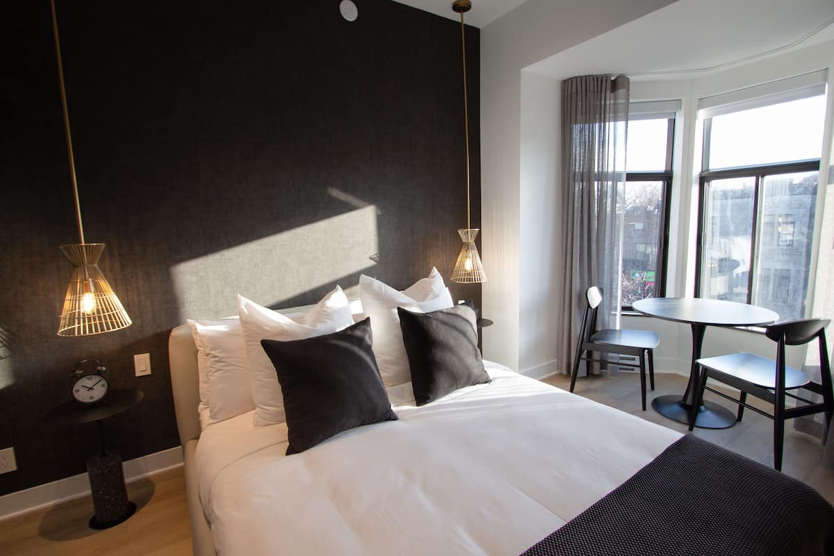 A hotel room with a large bed, two bedside tables, a desk with a chair, and a view of the city through large windows.