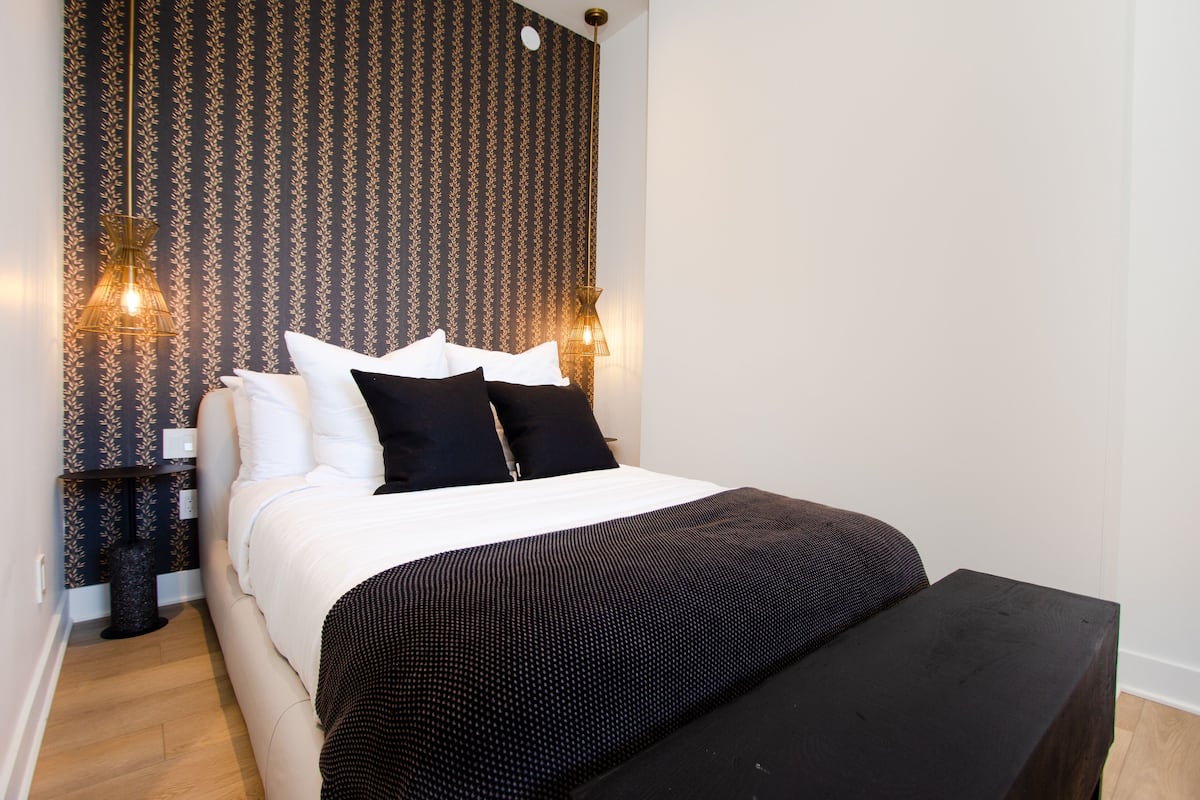 A neatly made bed with white and black bedding, two wall-mounted lamps, and a patterned wallpaper.