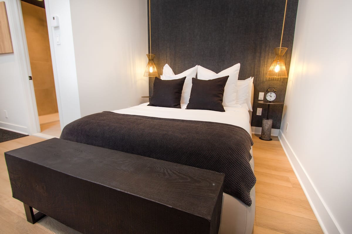 A modern bedroom with a large bed, bedside lamps, a wooden bench, and a clock on the nightstand.