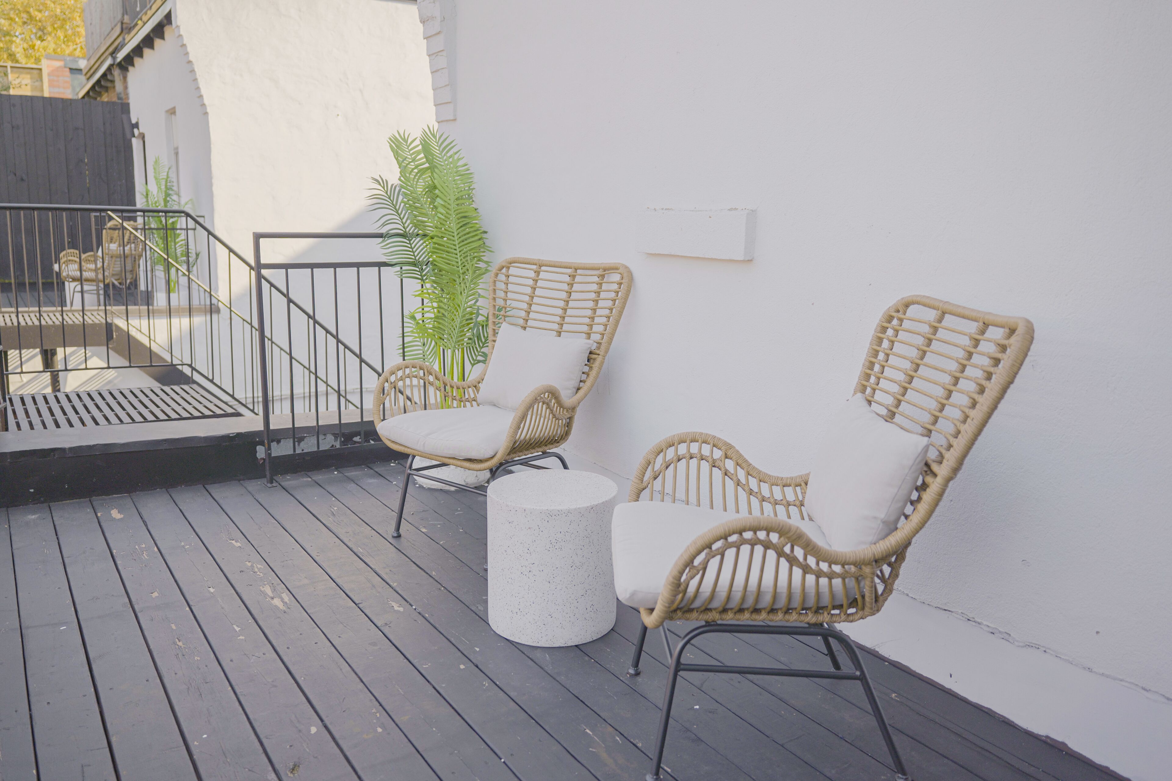 Una terraza con sillas de mimbre, una mesa redonda, paredes blancas y una barandilla metálica.