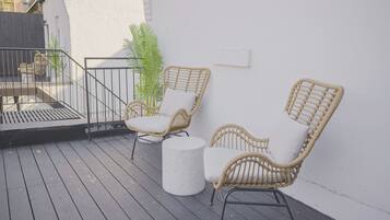 Una terraza con sillas de mimbre, una mesa redonda, paredes blancas y una barandilla metálica.