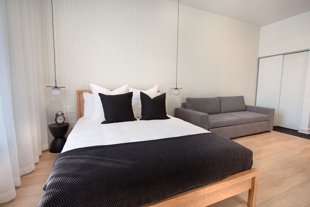 A modern bedroom with a large bed, a grey sofa, and a wooden bench.