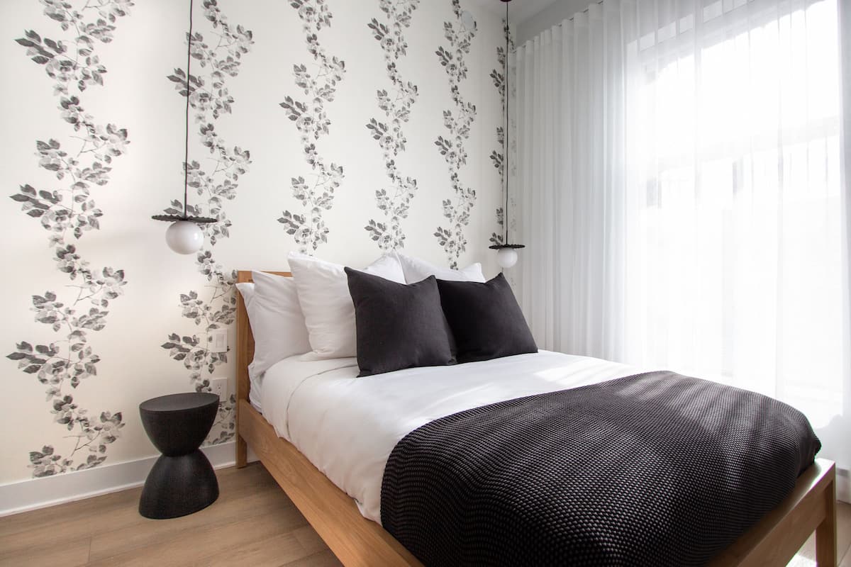 A neatly made bed with white and black bedding, a floral wallpaper pattern, and a round black side table.