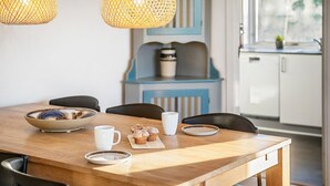 A wooden dining table with a bowl, a mug, and a plate with muffins. There are two chairs and a window in the background.