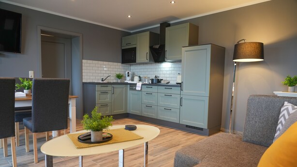 A modern kitchen with light-colored cabinets, a built-in oven, and a microwave. There is a dining area with a table and chairs, and a sofa with a cushion. - Aparthotel Rotheul (Föritztal)