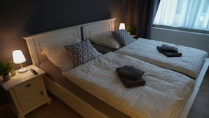 A double bed with white linens and two pillows, a bedside table with a lamp, and a window with curtains. - Aparthotel Rotheul (Föritztal)