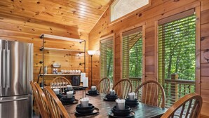 A wooden cabin interior with a dining table set for six, a kitchen area with a refrigerator, and large windows with a view of greenery. - Serene Wooded Family Mtn. Cabin- Great Location next to everything- Pet Friendly (Pigeon Forge)