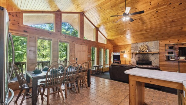 A spacious kitchen with a stone fireplace, wooden ceiling, and large windows overlooking a forested area. - Serene Wooded Family Mtn. Cabin- Great Location next to everything- Pet Friendly (Pigeon Forge)