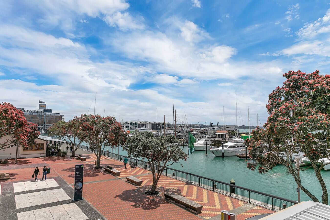 A marina with boats, a pedestrian walkway, and a building with a distinctive roof in the background.
