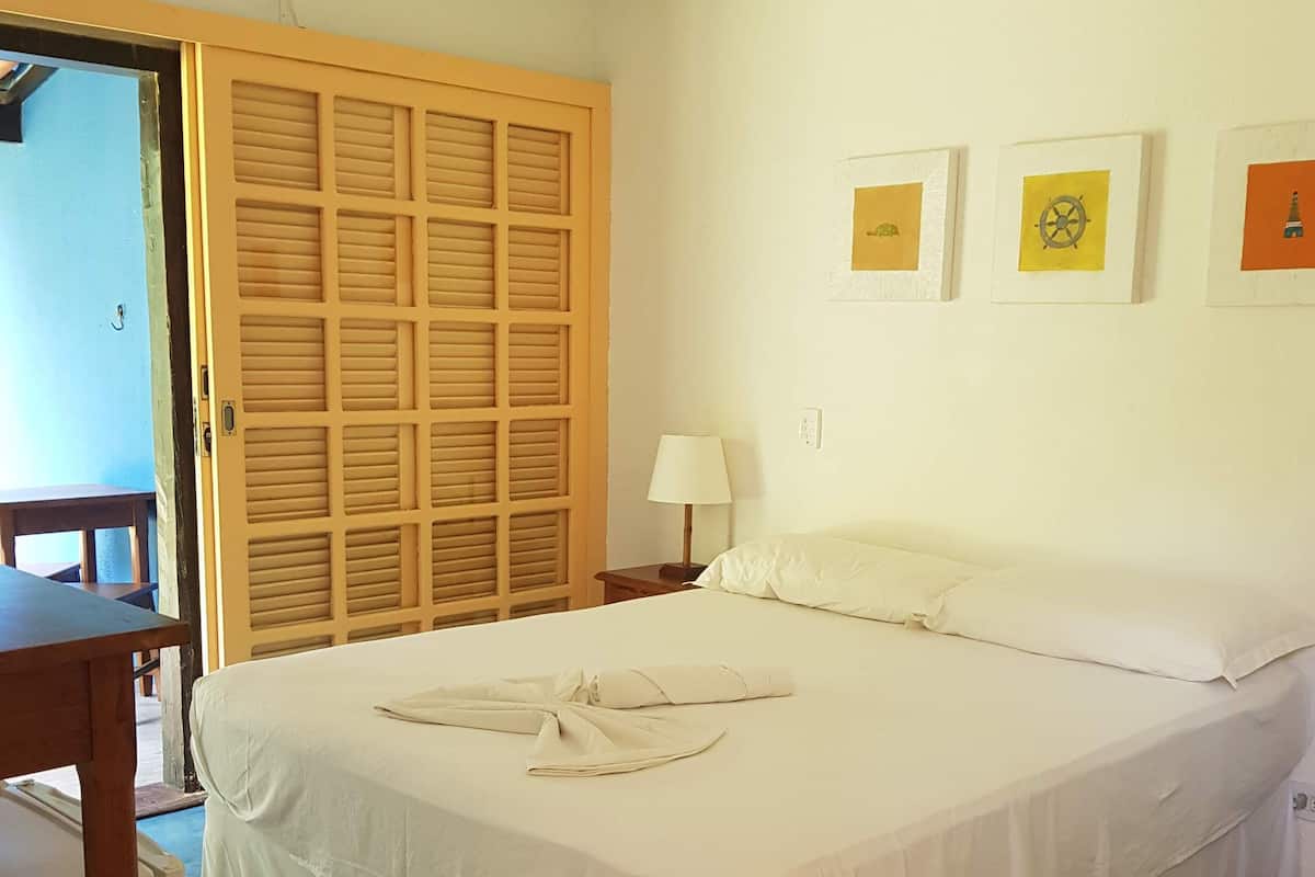 Una cama bien hecha con una colcha blanca y almohadas, una puerta de madera con diseño de rejilla, un ventilador de techo y una pequeña mesa con una silla.