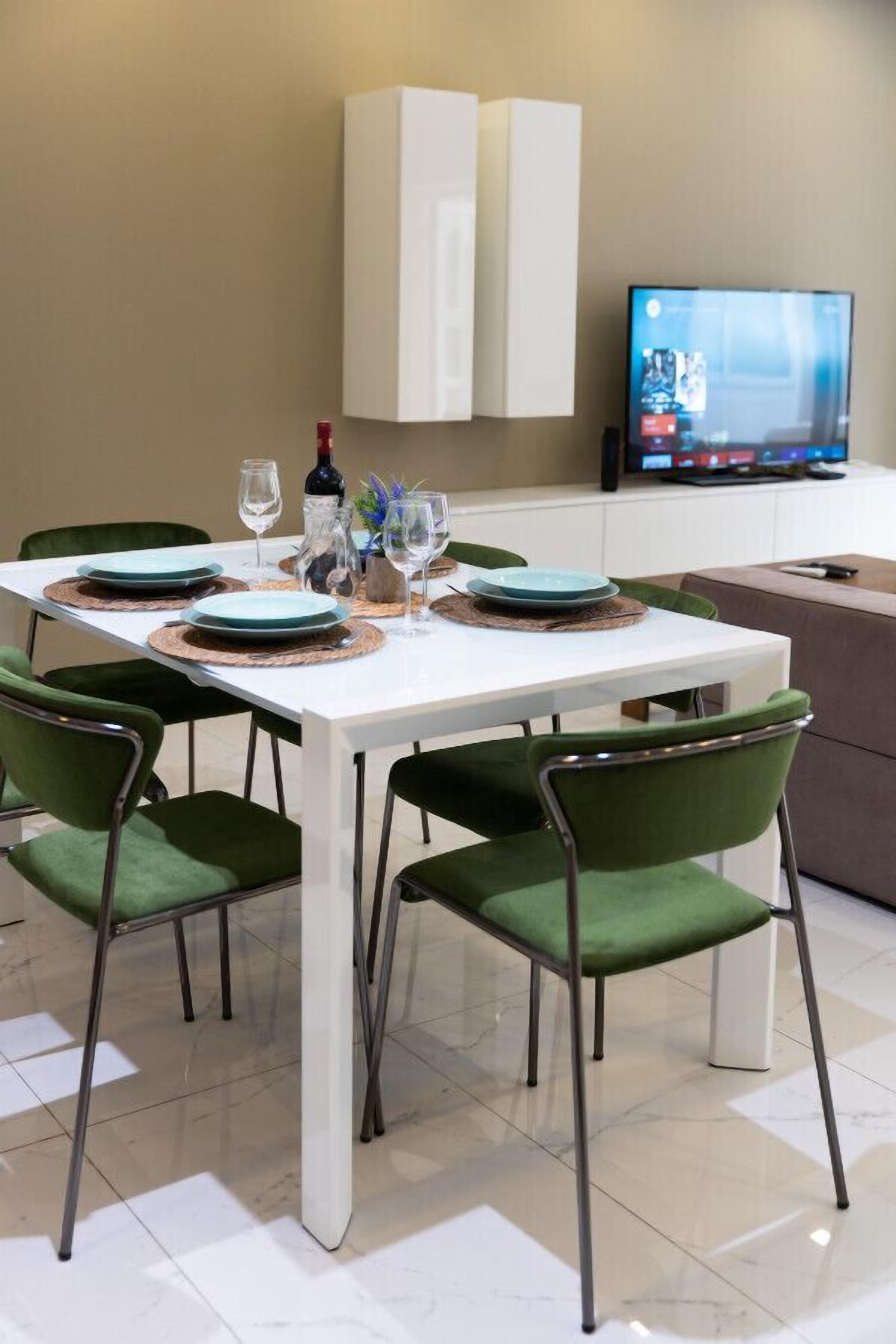 A dining area with a white table set for four, green upholstered chairs, and a television on the wall.