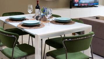 A dining area with a white table set for four, green upholstered chairs, and a television on the wall.