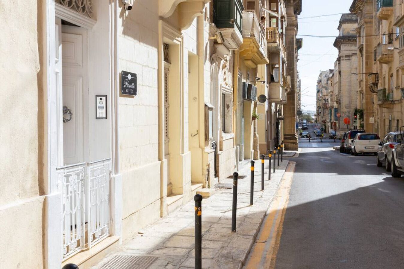 A row of traditional buildings with balconies, a street with cars, and bollards along the sidewalk.