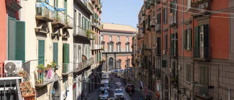 A narrow street lined with multi-story buildings, featuring balconies with plants and outdoor seating.