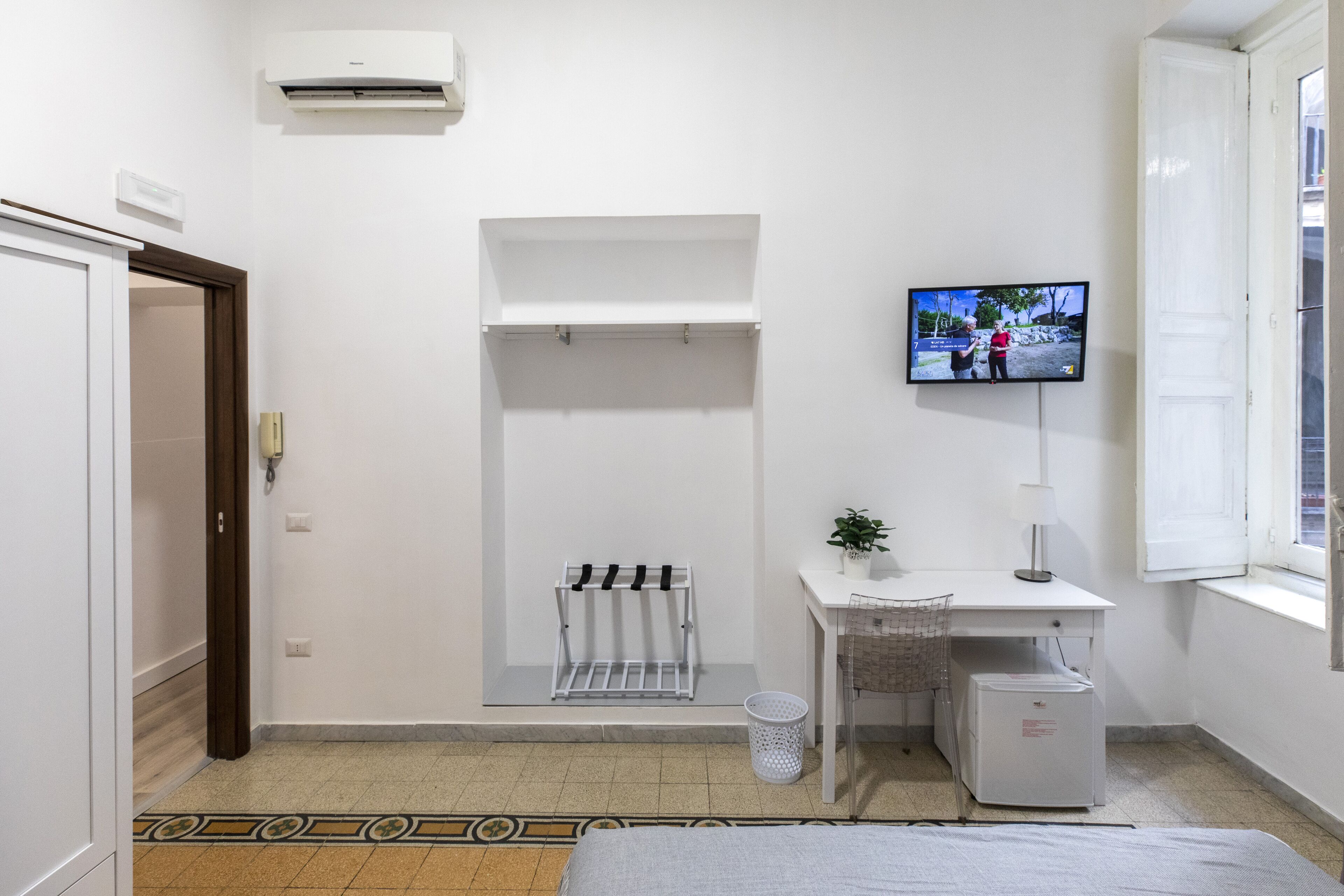 A small, clean room with a bed, a desk, a TV, a radiator, and a window.