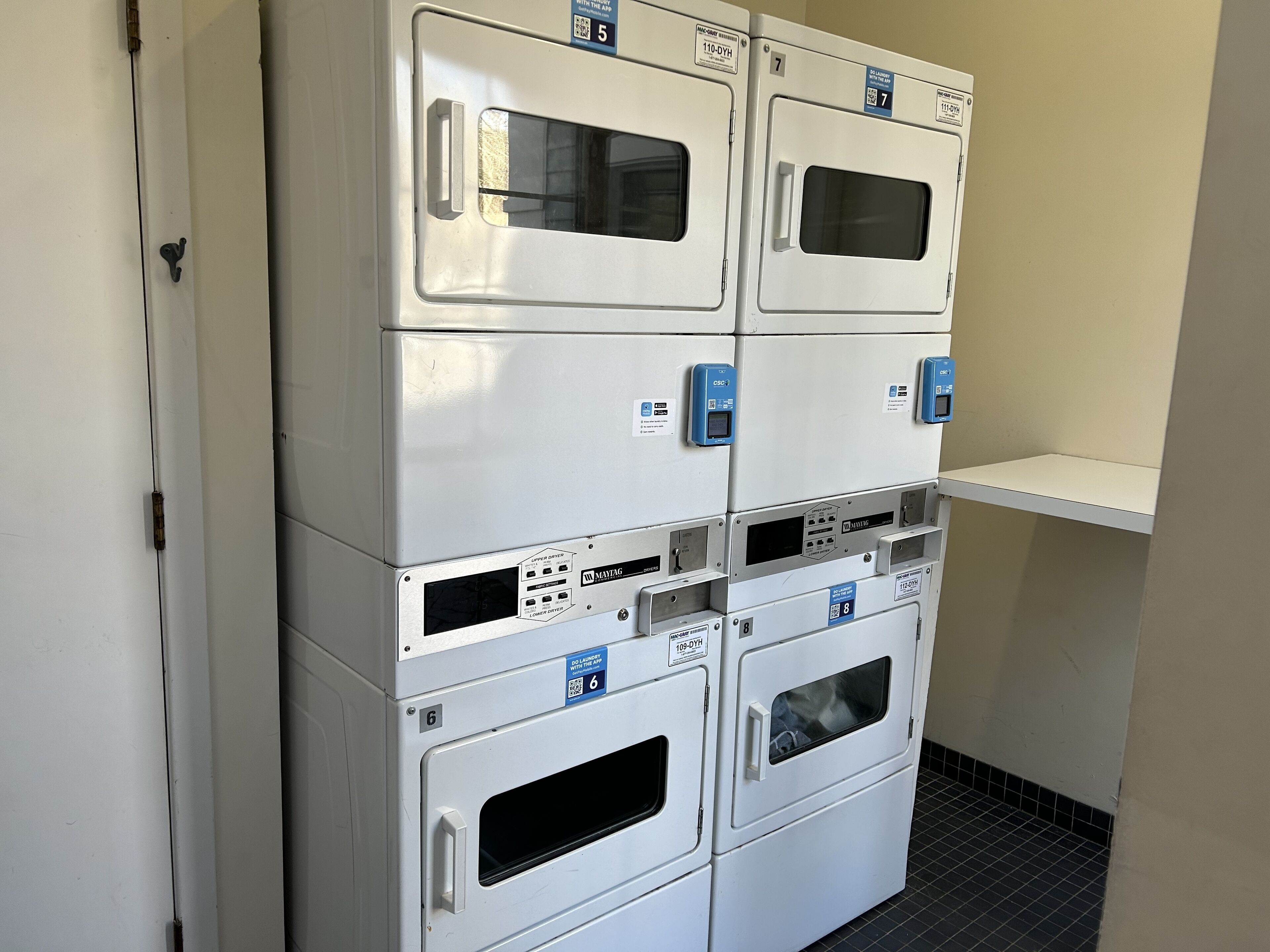 Une rangée de machines à laver blanches, dotées de panneaux de commande et de codes QR, est alignée contre un mur.