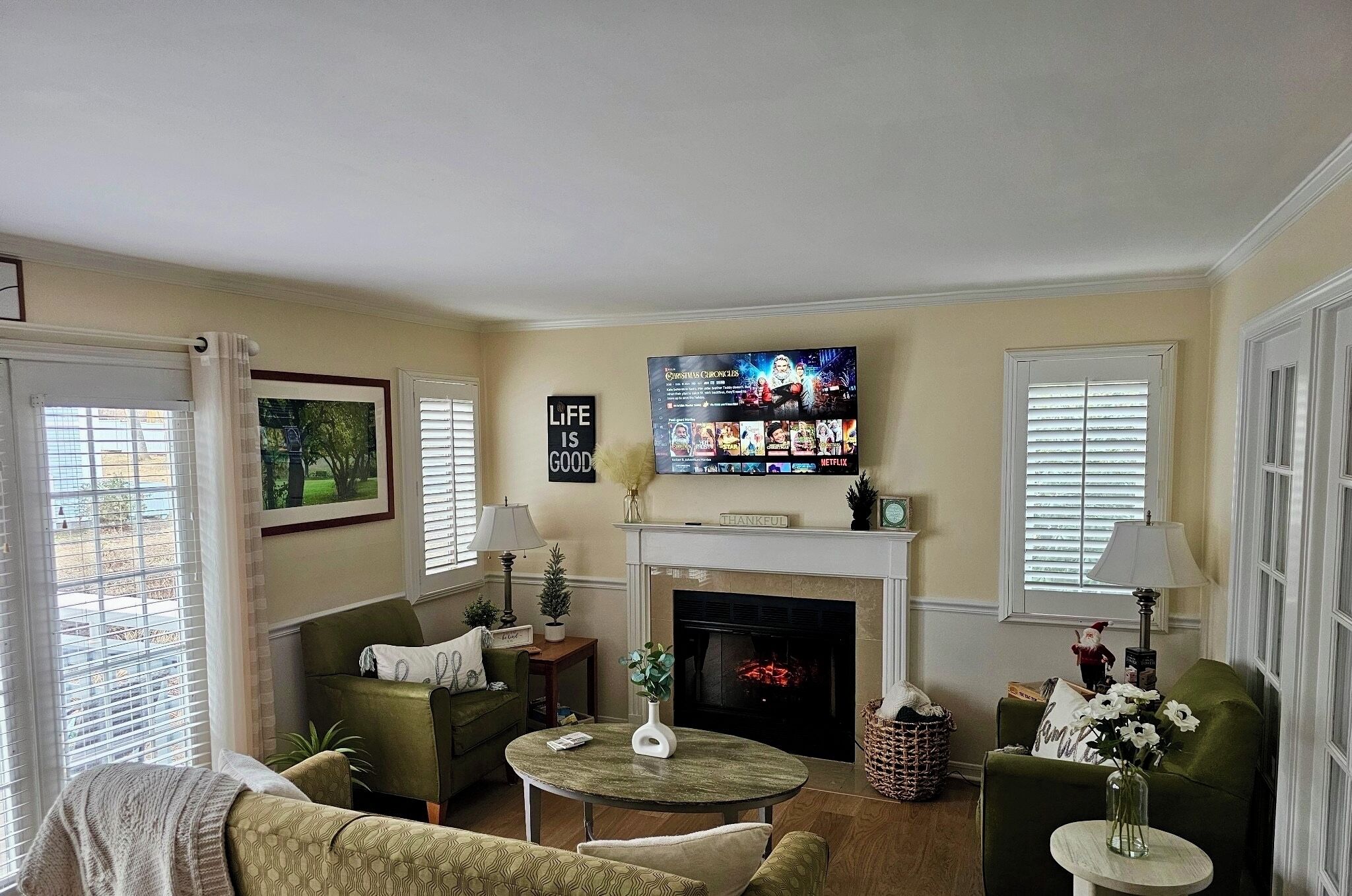 A living room with a fireplace, green sofas, a television, and a sign that says "Life is Good".