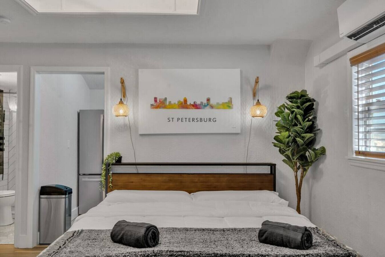 A bedroom with a bed, two wall-mounted lamps, a cityscape artwork, and a potted plant.