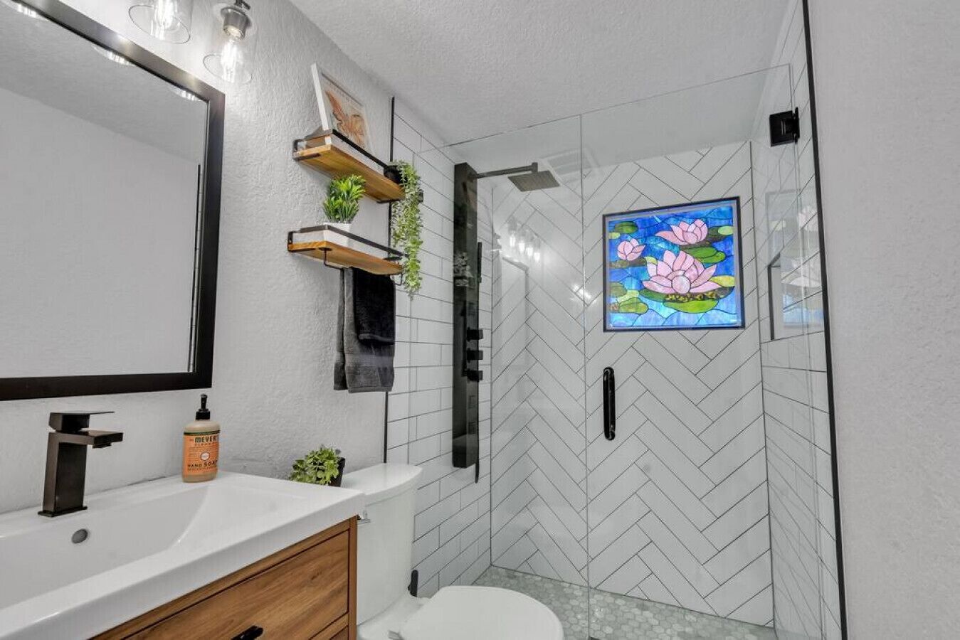 Um banheiro moderno com um espelho grande, pia branca, toucador de madeira, chuveiro com porta de vidro e parede decorativa com pintura de flor de lótus.