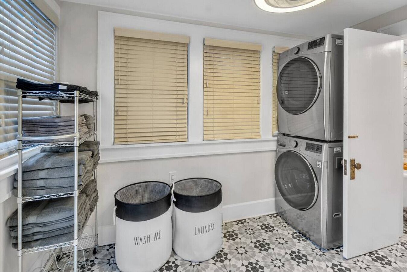 A laundry room with a stacked washer and dryer, a metal drying rack, and two laundry baskets labeled "WASH ME." and "LAUNDRY."
