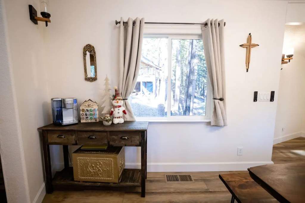 A room with a window, a wooden table, a small console table with decorative items, a mirror, and a cross on the wall.