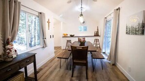 A dining area with a wooden table and chairs, a chandelier, and a view of the outdoors through large windows. - Cozy retreat near Dodge Ridge Ski Resort, and Pinecrest Lake with fast Wi-Fi  (Cold Springs)