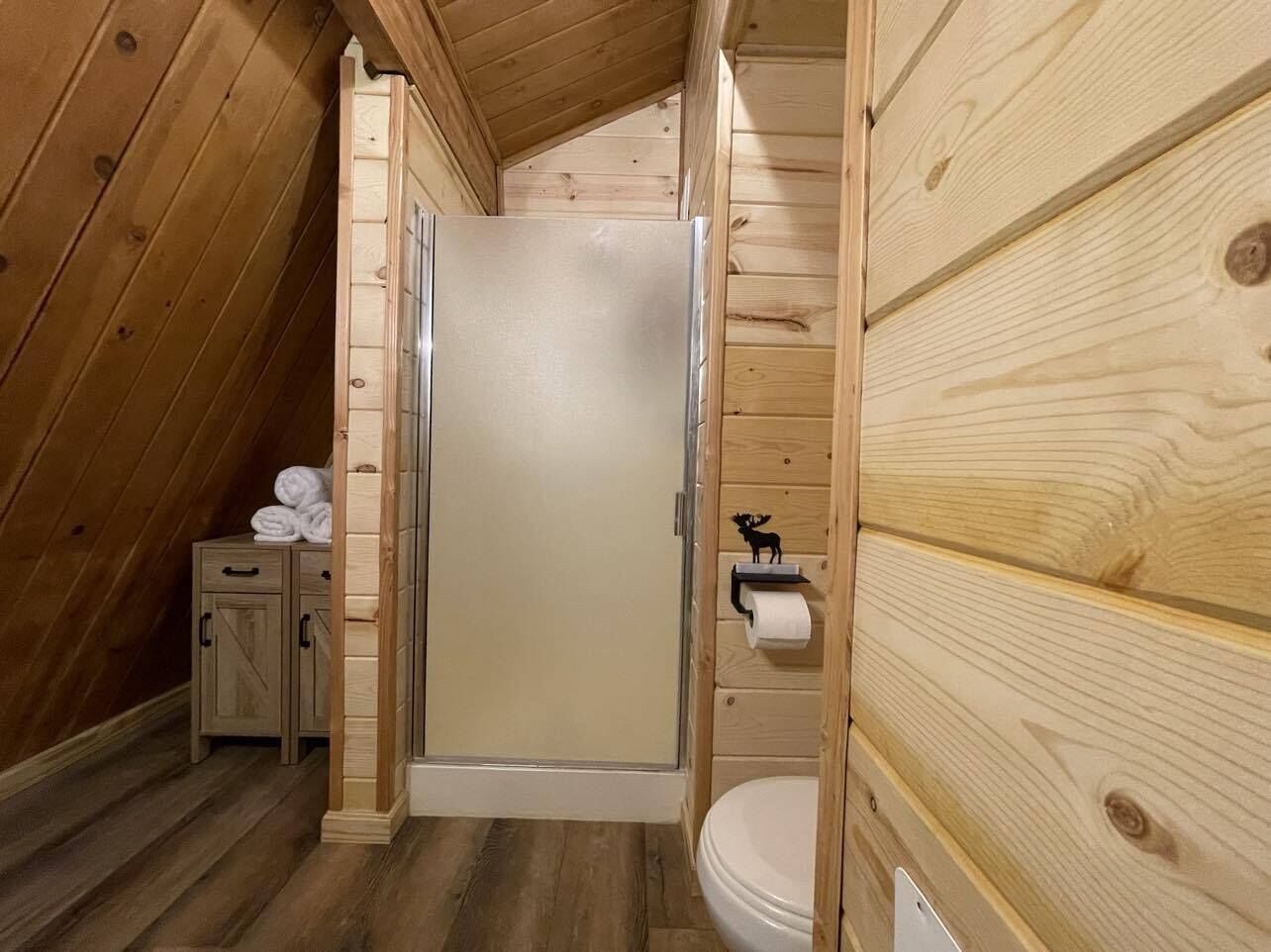 Un baño rústico de cabaña de madera, con ducha, inodoro y un mueble pequeño con toallas.