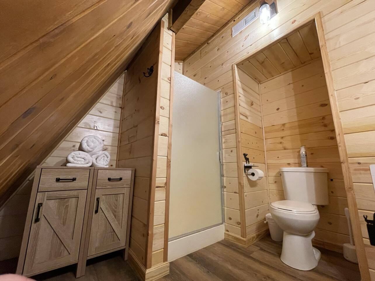 A wooden cabin with a lofted bathroom, including a toilet, a cabinet with towels, and a wooden ceiling.