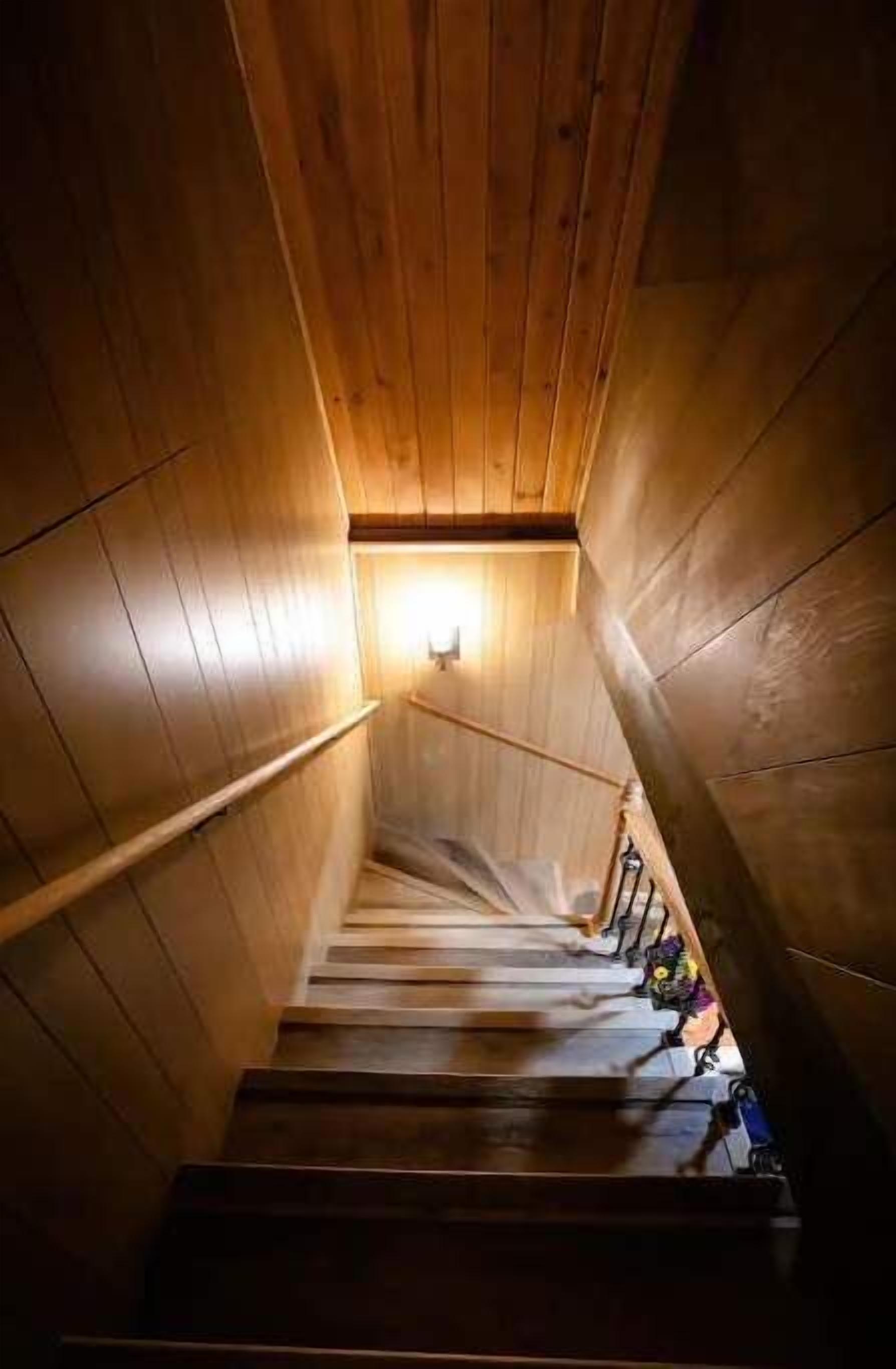 Un escalier en bois, doté d’une rampe, qui monte vers une source de lumière située en haut.