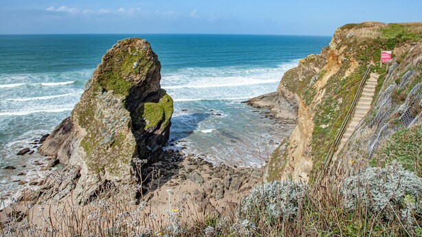 Un paysage côtier caractérisé par une formation rocheuse imposante, un escalier menant à une falaise et un panneau indiquant qu’il faut faire preuve de prudence.