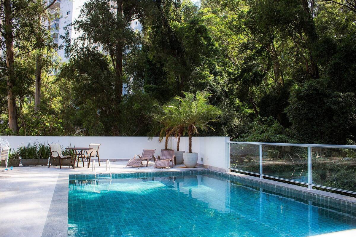 Une piscine entourée d’arbres, avec des chaises longues et un mur blanc.