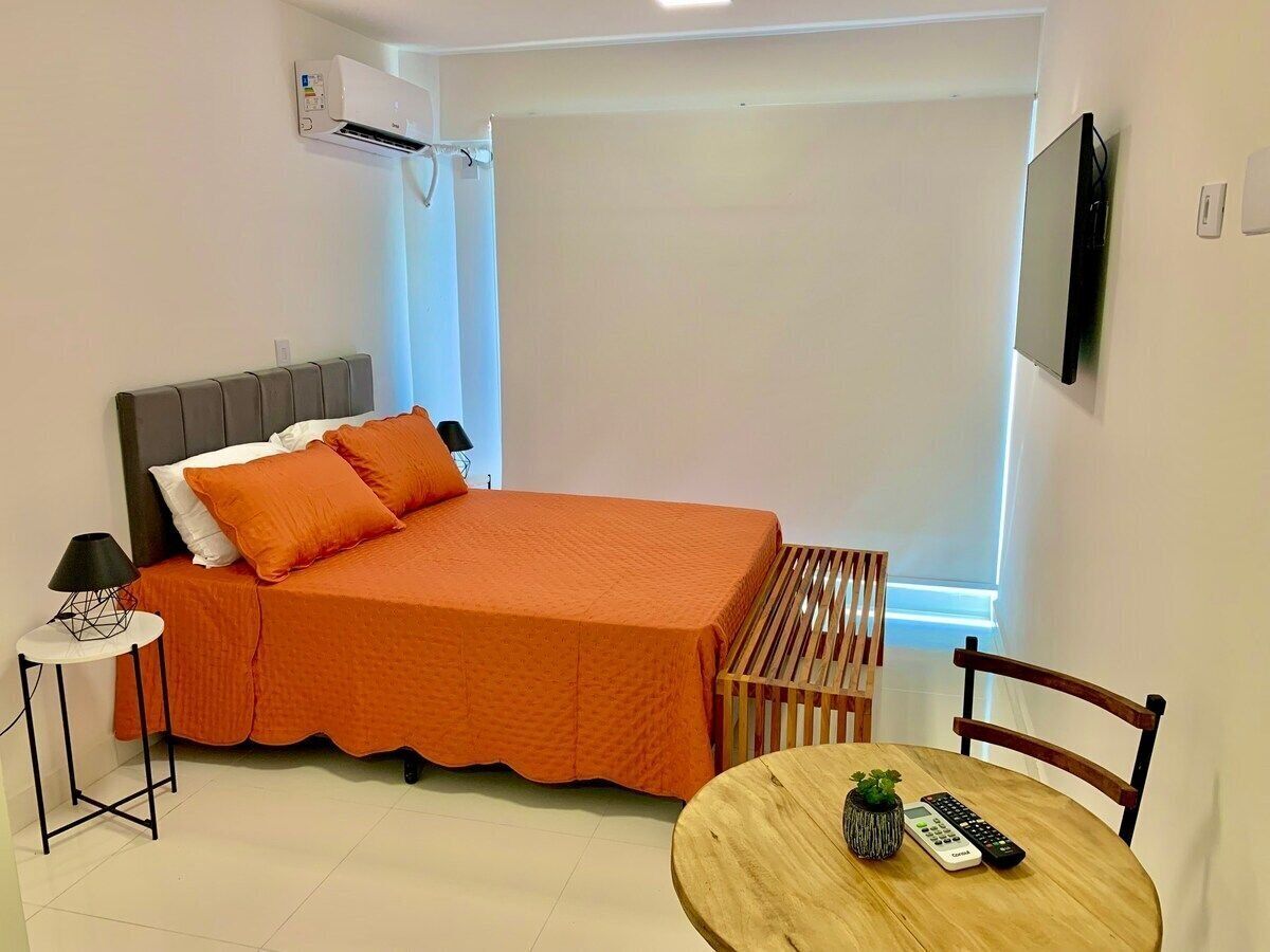 A bedroom with a bed, a bedside table with a lamp, a wooden chair, a small table with a plant and remote controls, and a mounted TV.