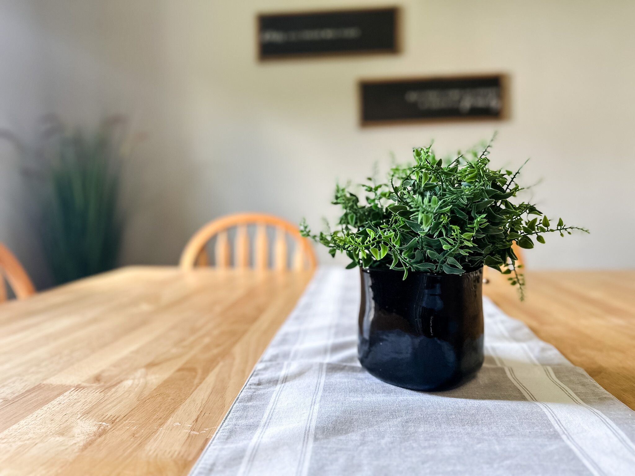 Uma mesa de madeira com um vaso escuro de plantas verdes sobre um pano de mesa branco.