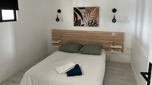 Une chambre avec une tête de lit en bois, un lit avec une literie blanche et verte, deux lampes de lecture fixées au mur et un climatiseur fixé au mur.