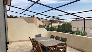 Une terrasse extérieure couverte, avec une table et des chaises, un sol carrelé et une vue sur les bâtiments et les arbres en arrière-plan.