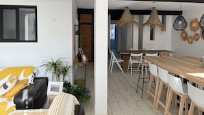 Un intérieur moderne comprenant un coin repas avec une table en bois et des chaises blanches, un coin salon avec un canapé et des coussins, et un couloir agrémenté d’un miroir et de luminaires suspendus.